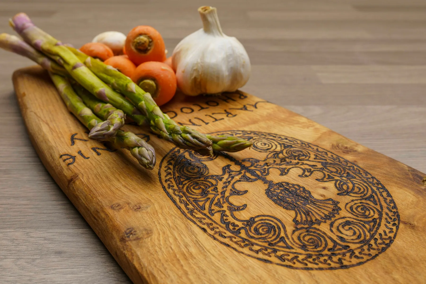 A wooden cutting board engraved with an ornate circular medallion, with fresh asparagus, carrots, and garlic resting on it